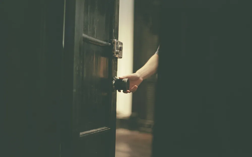 Puerta que se abre hacia un interior oscuro, con una mano en la manilla